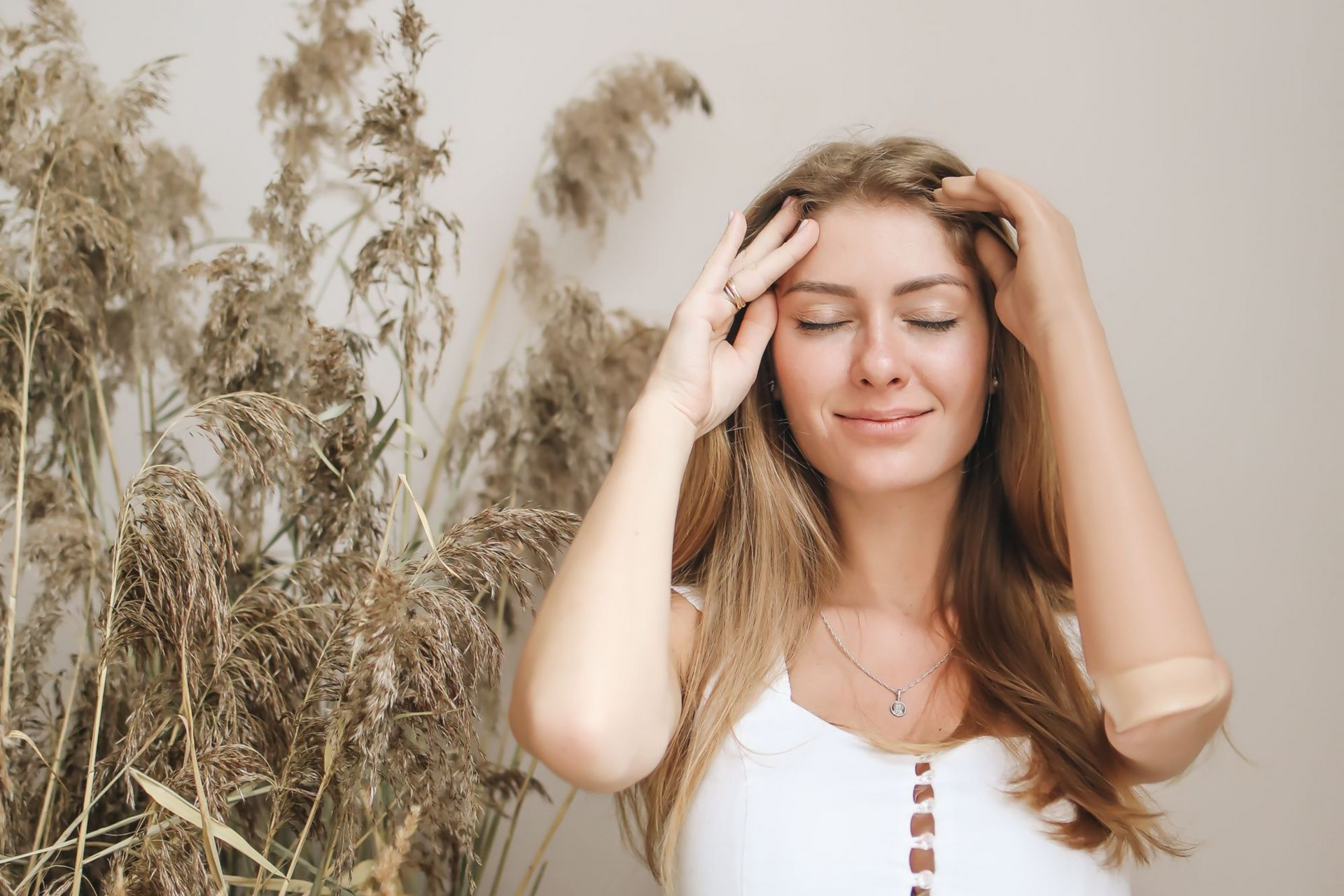 Woman standing outdoors among tall grass, eyes closed, touching her hair in soft sunlight.
