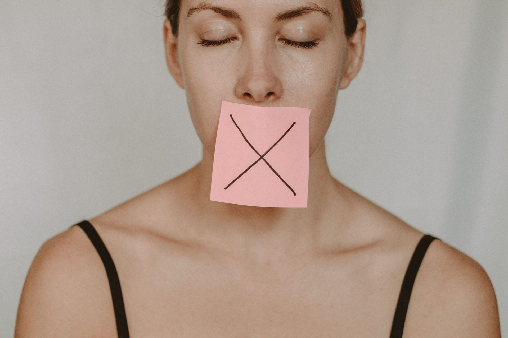Close-up of a woman with eyes closed, holding a pink note over her mouth with an X drawn on it.