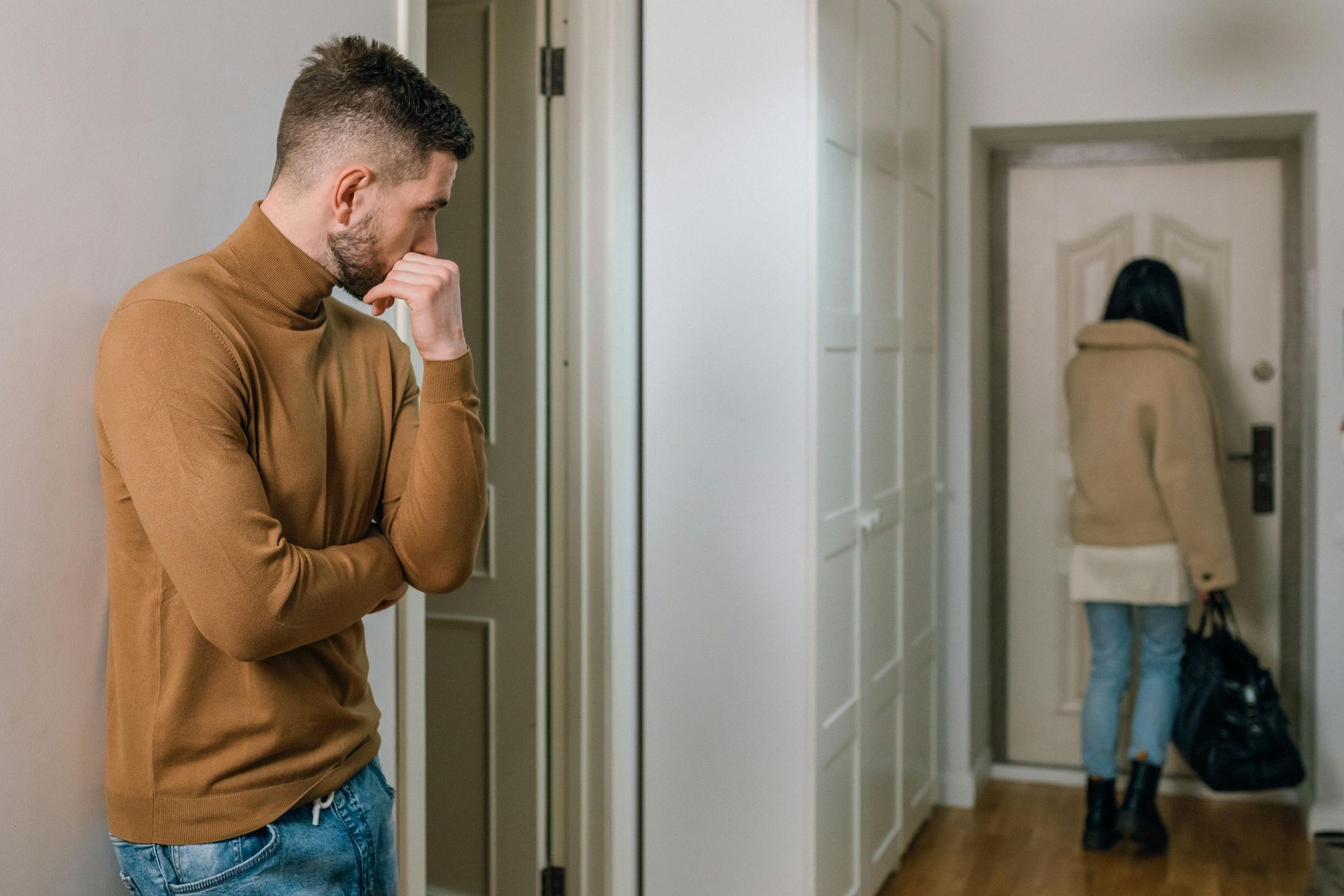 Man watches woman near a door; he is in a brown sweater, she has a coat, and a bag.