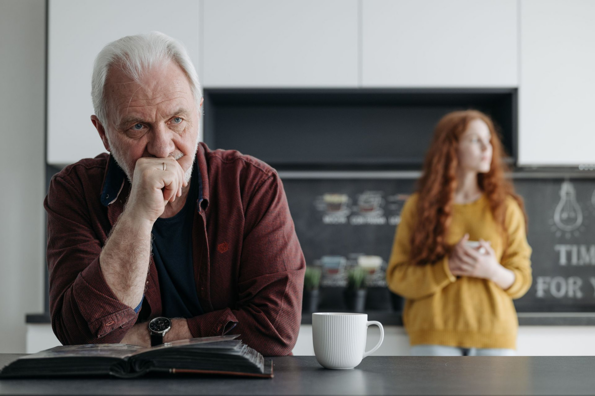 Man sitting at a table with an open book and mug, looking thoughtful; a woman stands blurred in the background.