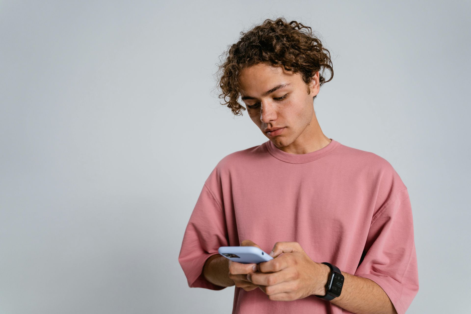 Young person with curly hair, wearing a pink shirt, looks down at their phone in their hands.
