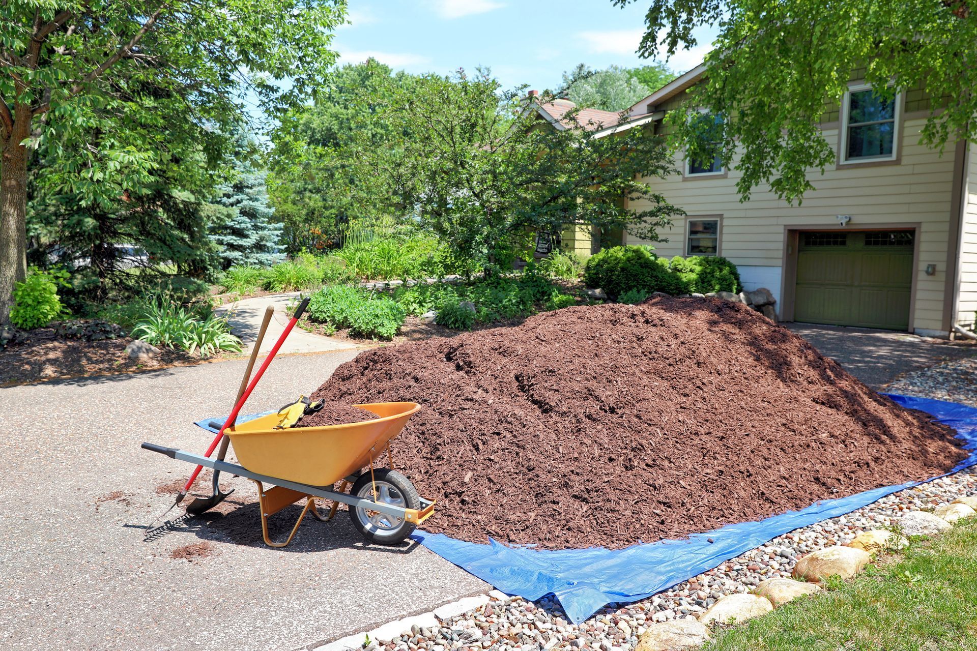 Large pile of brown mulch on blue tarp in a driveway with a wheelbarrow and tools. House and trees in background.
