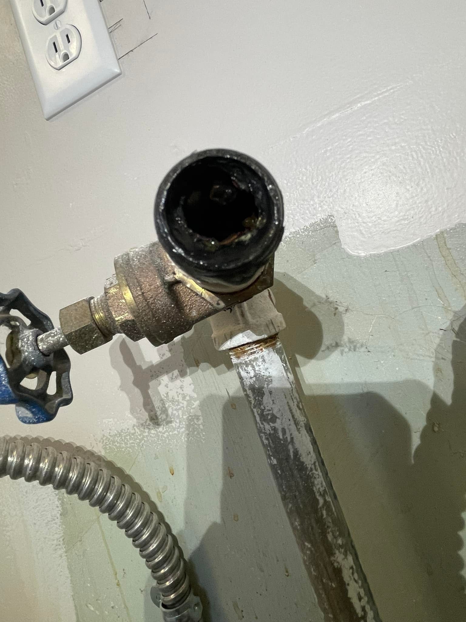 Close-up of a corroded black pipe fitting attached to a brass valve on a light gray wall.