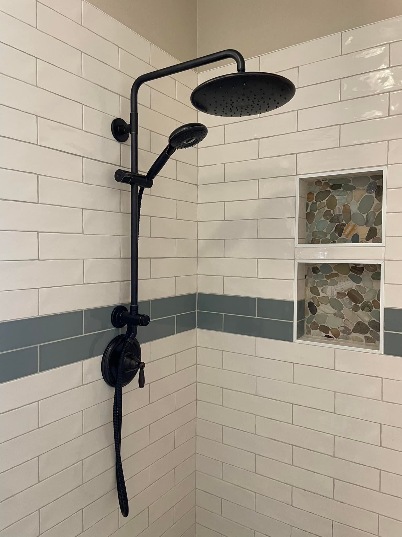 Black shower fixture with overhead rain showerhead and handheld sprayer, mounted on white tile with decorative insets.