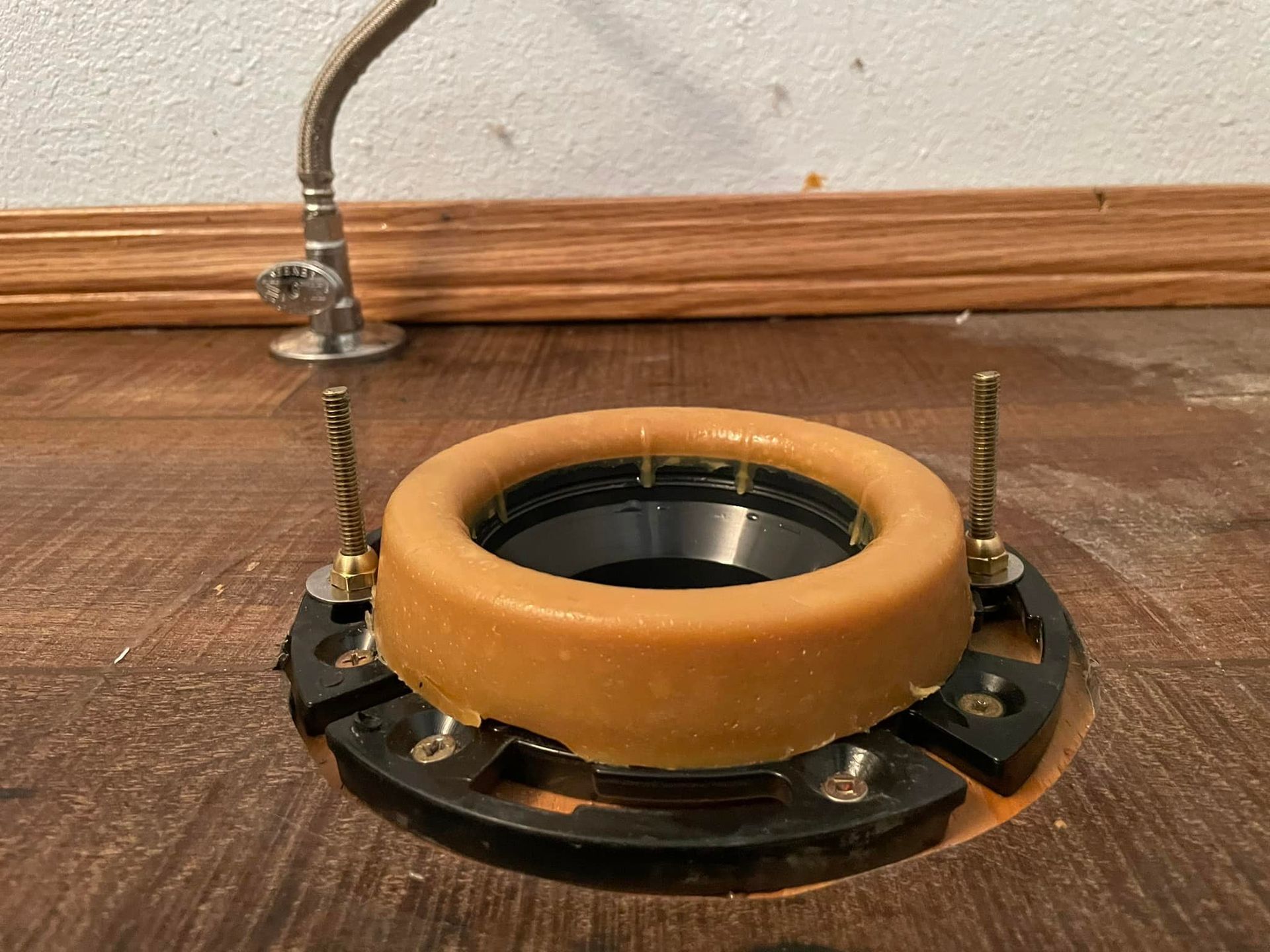 Toilet flange with wax ring installed, ready for toilet. Brass bolts protrude. Brown floor.