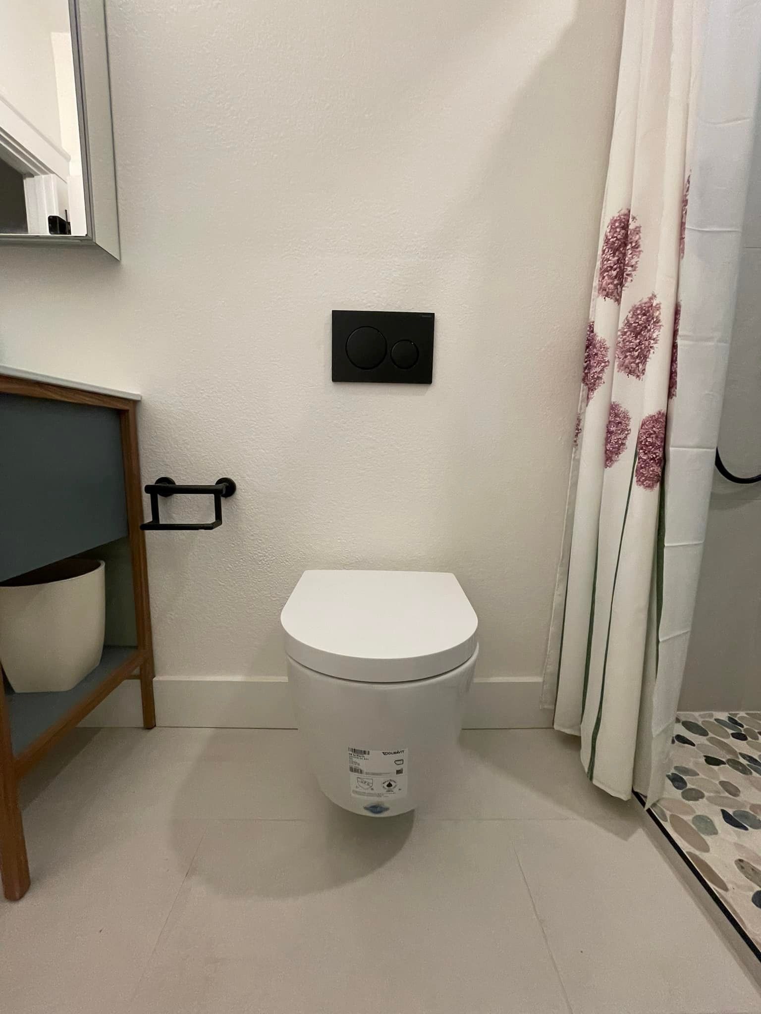 White bathroom with wall-mounted toilet, black flush plate, and shower curtain with floral pattern.