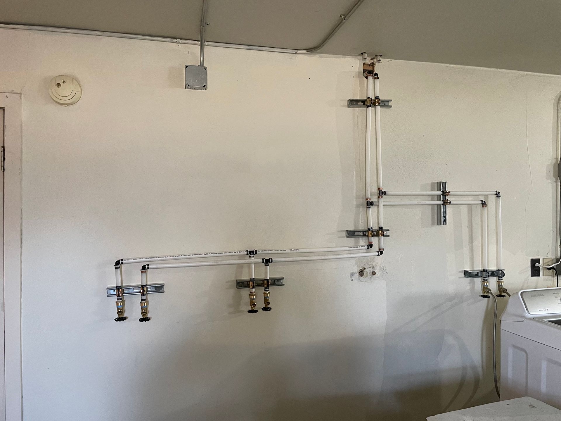 Piping and fixtures mounted on a white wall. The pipes run horizontally and vertically, with several valves and fixtures installed.