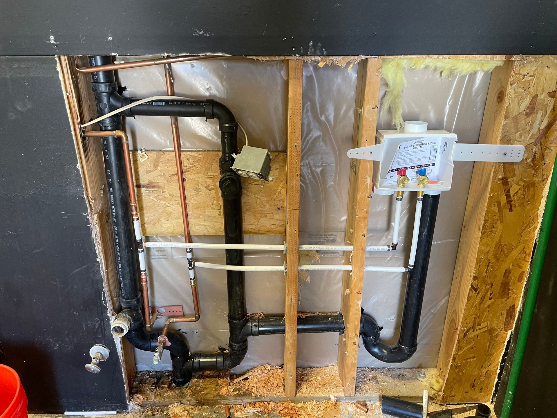 Plumbing rough-in inside a wall cavity. Black drain pipes, copper water lines, and electrical box are visible.