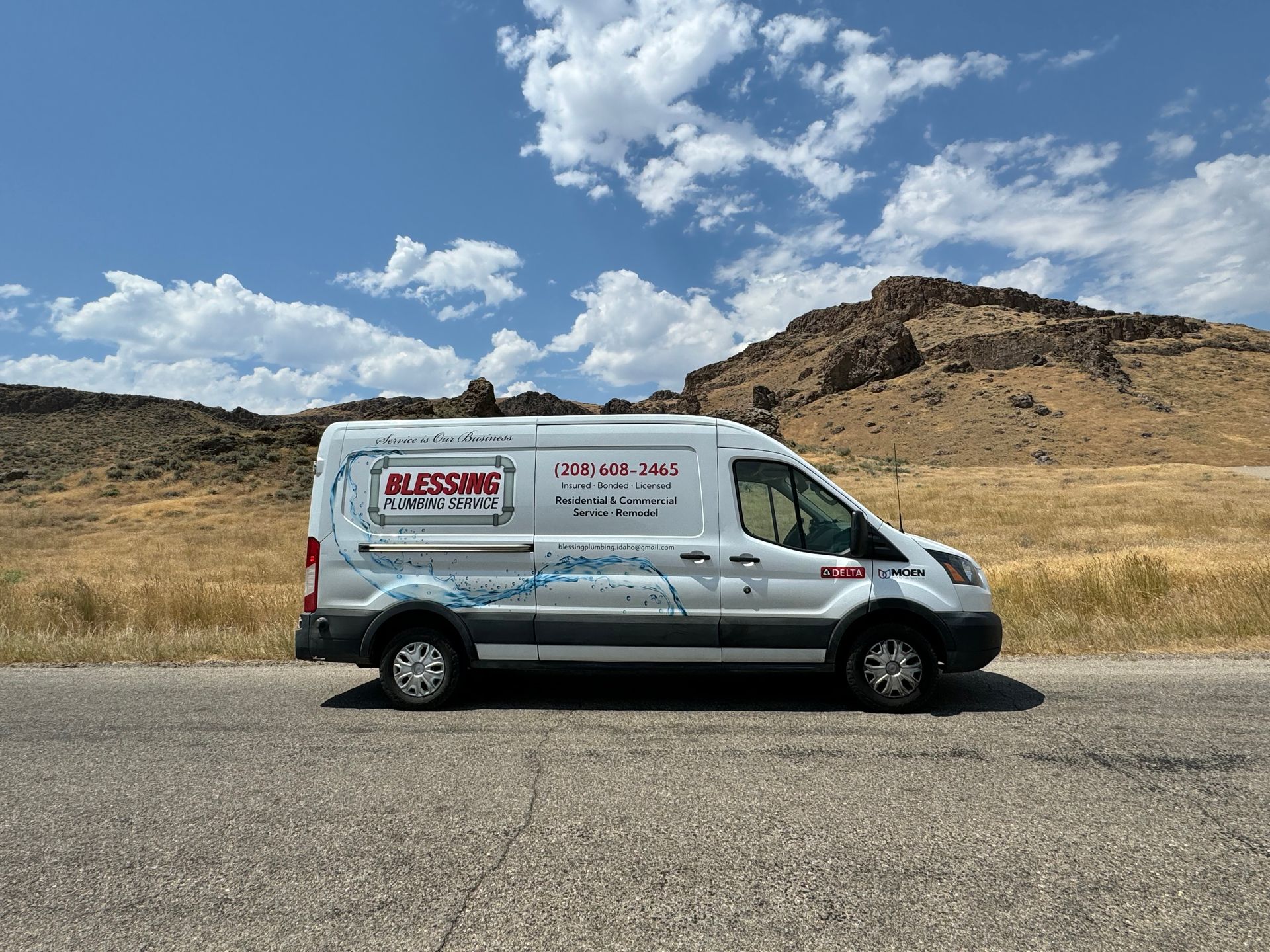 White service van with business logo parked on roadside, mountains and blue sky in background.