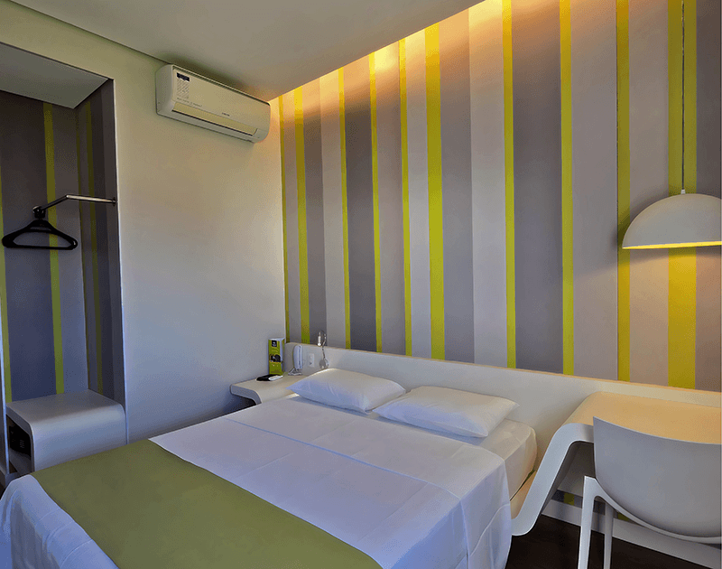 Um quarto de hotel com cama, escrivaninha branca, cadeira, papel de parede listrado em amarelo e cinza e ar-condicionado de parede.