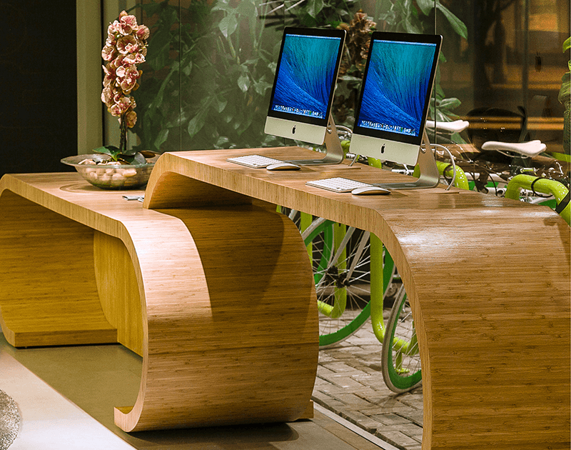 Uma mesa moderna e curva de bambu com dois computadores, posicionada em frente a uma vitrine interna com bicicletas e uma orquídea.