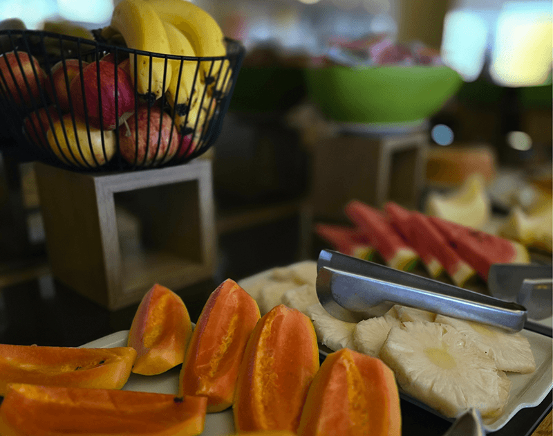 Uma apresentação de buffet com uma cesta de bananas e maçãs sobre fatias de mamão e abacaxi em uma bandeja de servir.
