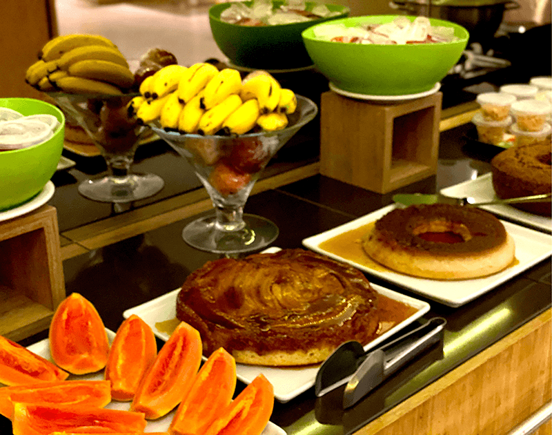 Um buffet de café da manhã com tigelas de frutas, copos de bananas e fatias de mamão, e travessas de bolos de pudim de caramelo.