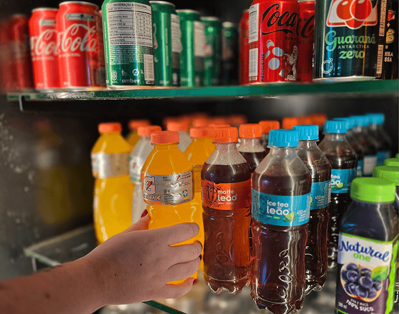 Uma mão alcança uma garrafa de refrigerante laranja brilhante na prateleira da geladeira de uma loja, repleta de diversas bebidas e latas.