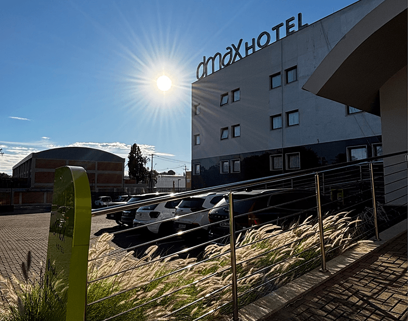 O edifício do Hotel Dmax sob um sol brilhante, com um estacionamento e grama alta e esparsa em primeiro plano.
