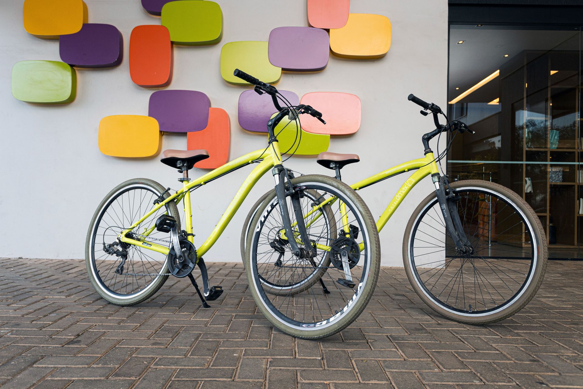 Duas bicicletas amarelas estacionadas em um pátio de tijolos estampados, em frente a um muro decorado com blocos retangulares coloridos.