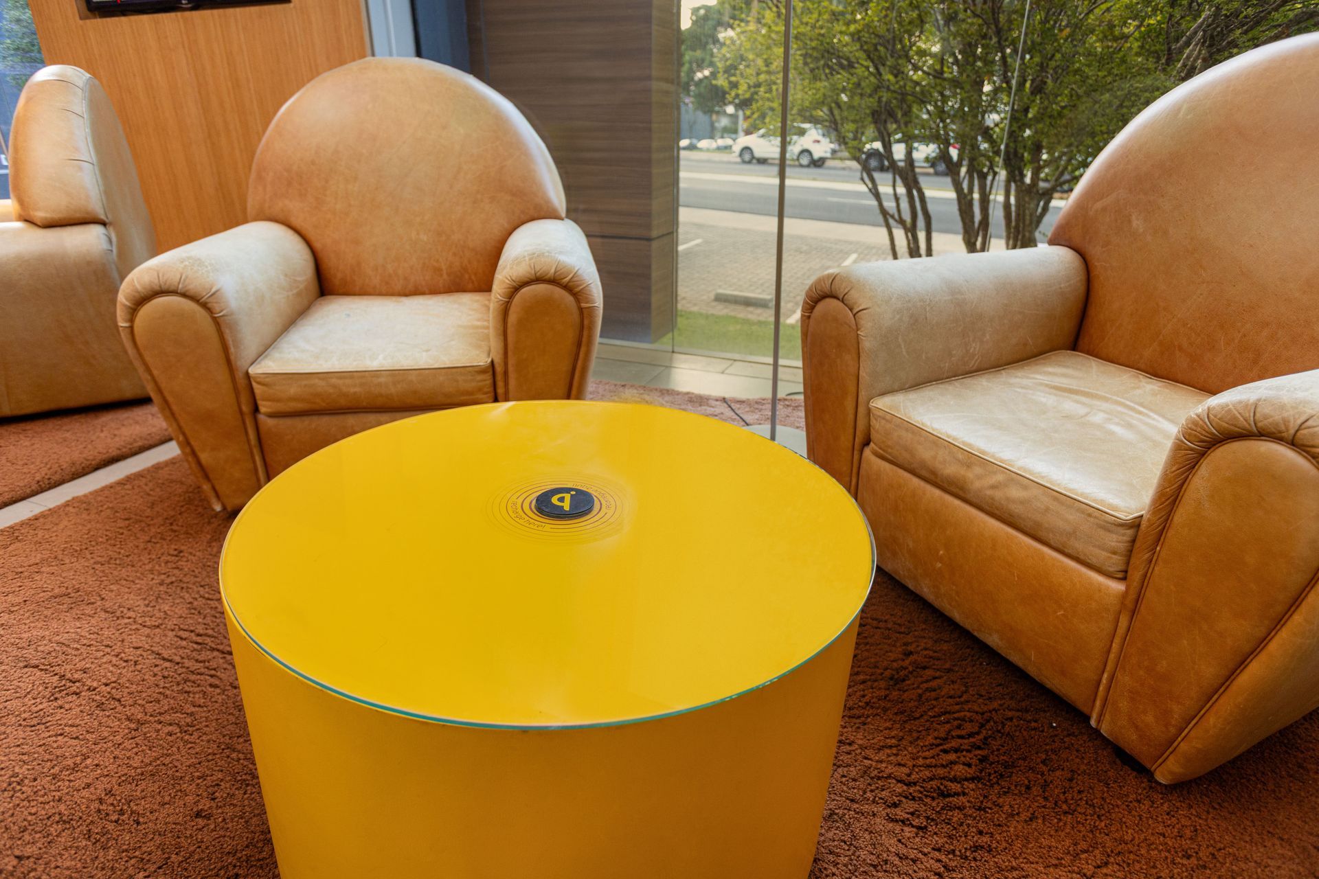 Duas poltronas bege ficam frente a frente em frente a uma mesa de pedestal circular amarela brilhante, sobre um tapete marrom perto de uma janela.