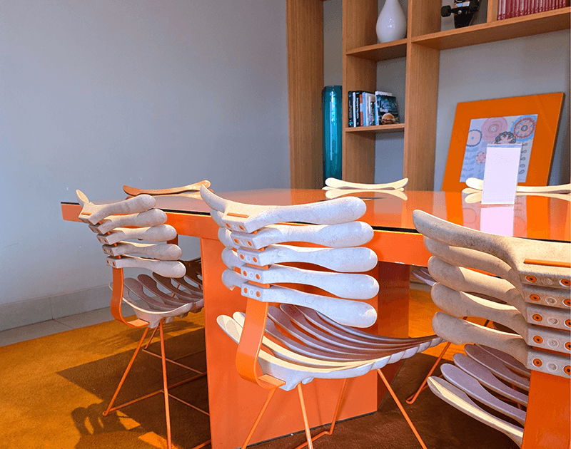 Sala de jantar moderna com uma mesa laranja vibrante e cadeiras exclusivas com encostos e estruturas de madeira branca em formato de costelas.