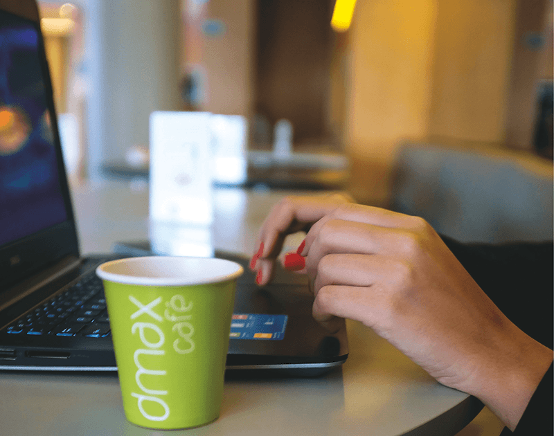 Uma mão com unhas pintadas de vermelho trabalha em um laptop ao lado de um copo de papel verde do café dmax, sobre uma mesa.