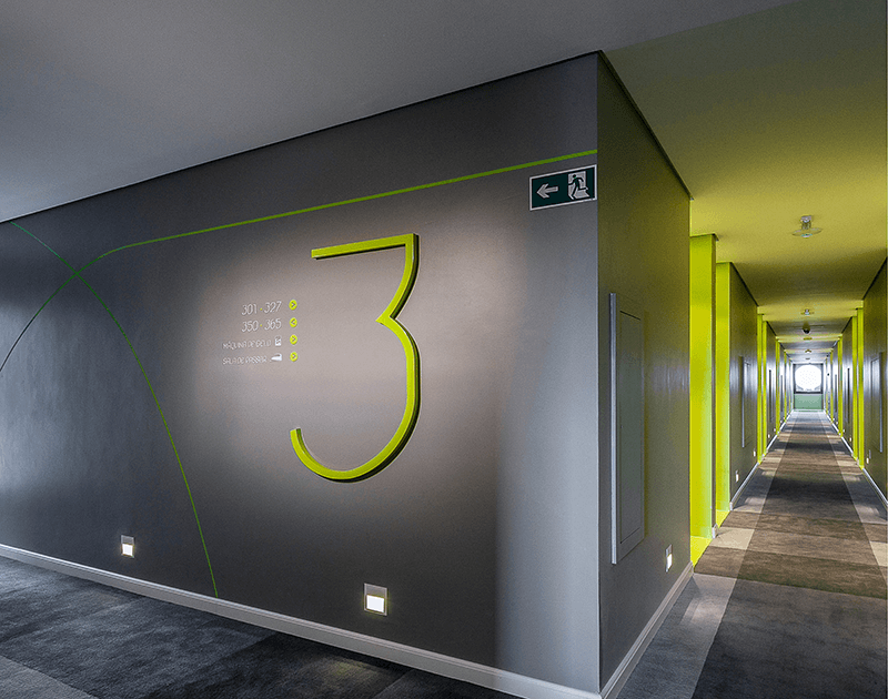 Um corredor com um grande número 3 verde neon em uma parede cinza escura, que leva a um longo corredor com detalhes em amarelo vibrante.