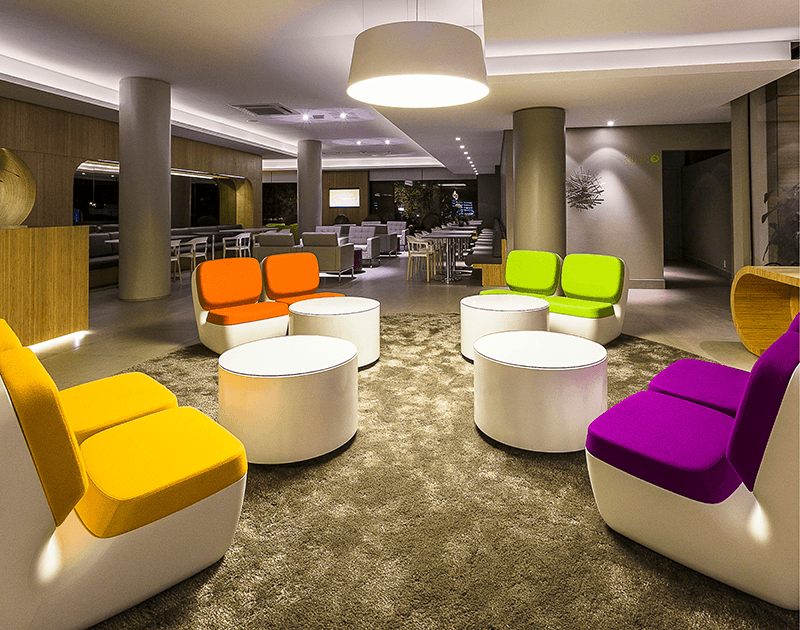 Um lobby de hotel moderno com assentos modulares coloridos, mesas redondas brancas e um tapete macio sob iluminação suave.