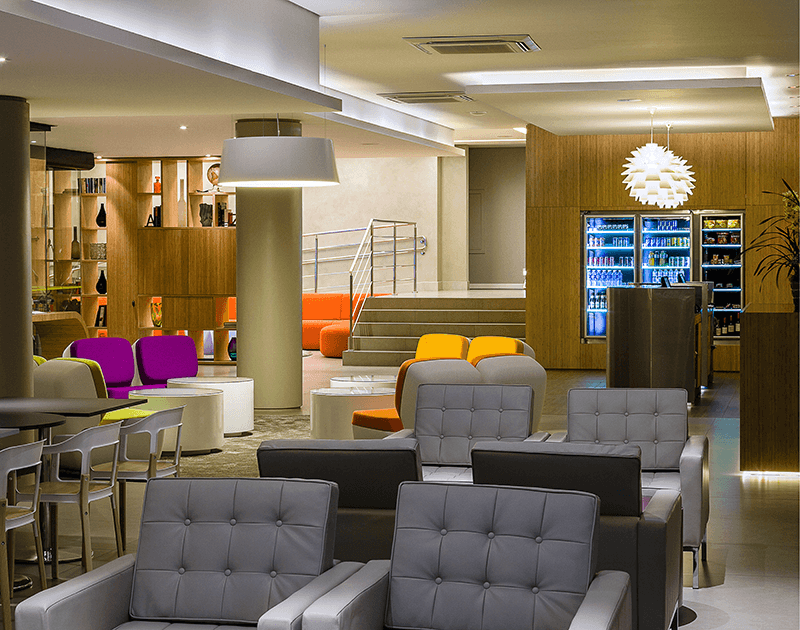 Um lounge de hotel moderno com poltronas estofadas cinza, cadeiras com detalhes em roxo, assentos laranja e uma máquina de venda automática iluminada.