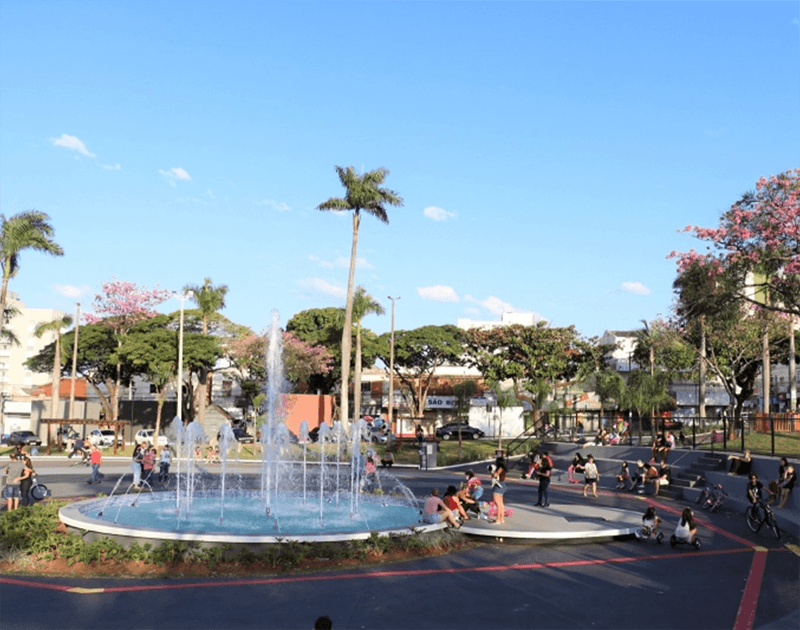 Uma fonte circular jorra água em uma praça pública ensolarada, cercada por árvores, pedestres e áreas pavimentadas para caminhada.