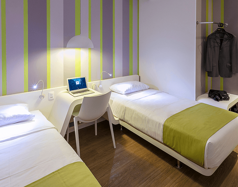 Um quarto de hotel moderno com duas camas de solteiro, uma escrivaninha, uma cadeira branca com um laptop e papel de parede listrado em tons de roxo e verde-limão.