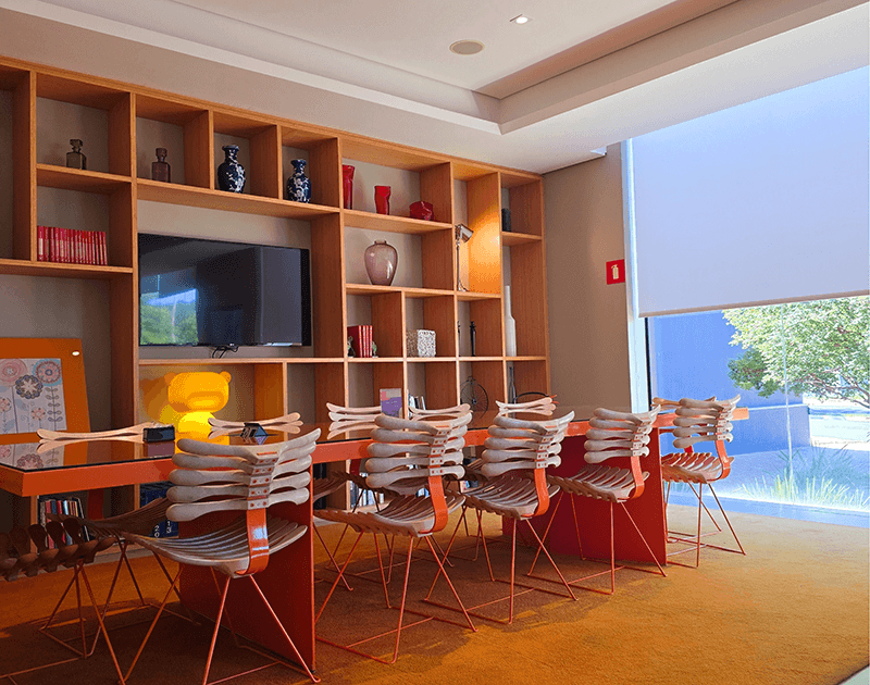 Uma sala de conferências com uma longa mesa laranja e cadeiras brancas de design abstrato em frente a uma estante de madeira que vai do chão ao teto.