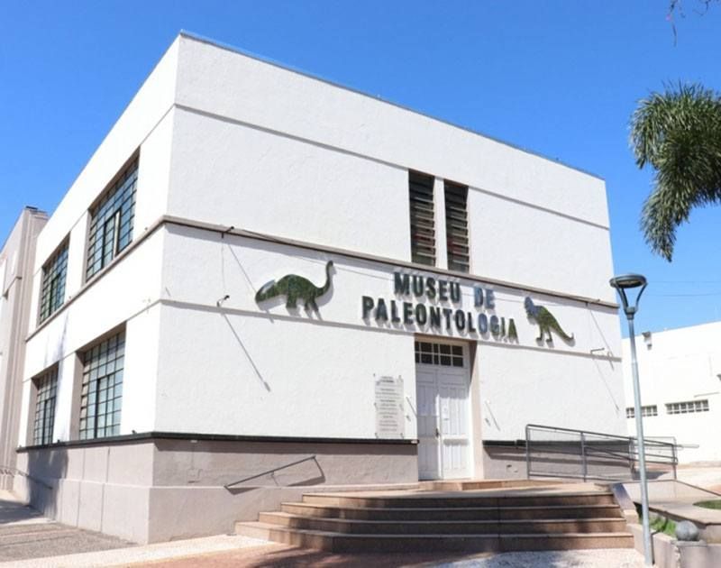 O edifício branco de dois andares do Museu de Paleontologia, com silhuetas de dinossauros na fachada frontal, sob um céu azul claro.
