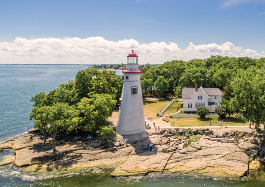 White lighthouse on a rocky shoreline beside a white house, with blue water and green trees under a cloudy sky