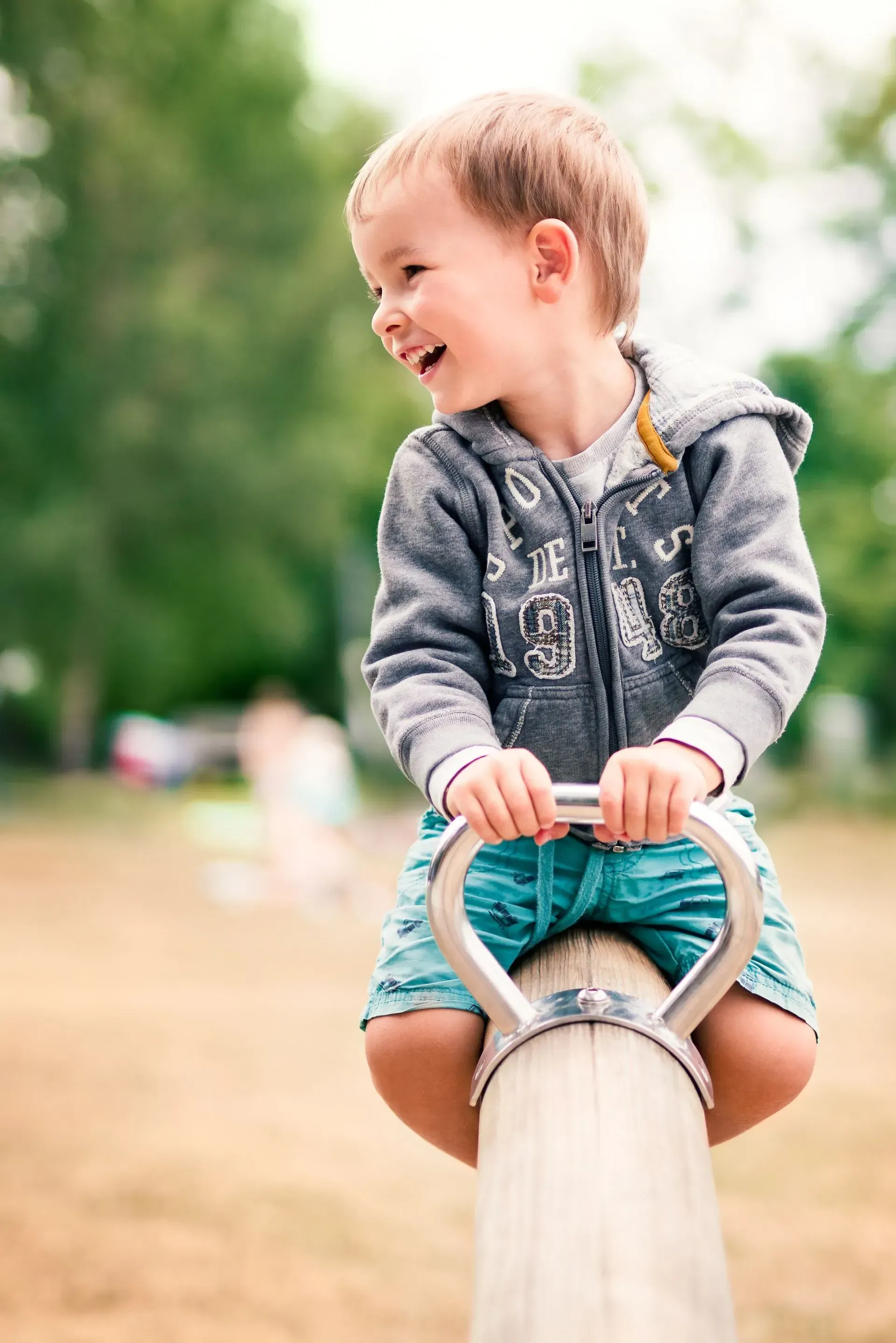 Child smiling while seated on a wooden playground balance beam, holding a turquoise ring outdoors.