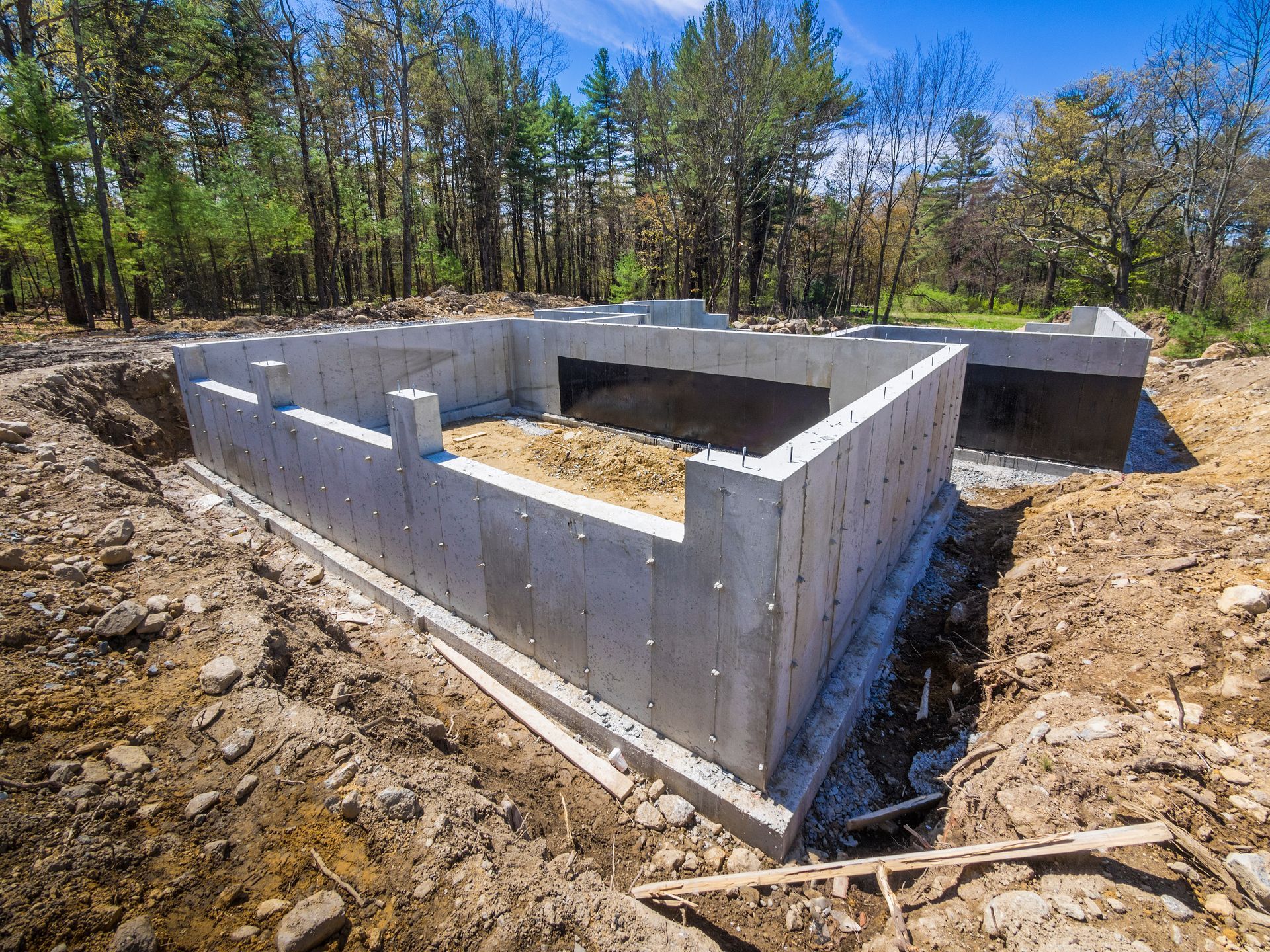 Murs de fondation en béton en construction sur un terrain boisé, avec de la terre et un ciel bleu.