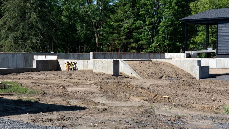 Chantier de construction avec murs de fondation en béton et tas de terre à côté d'une lisière de forêt