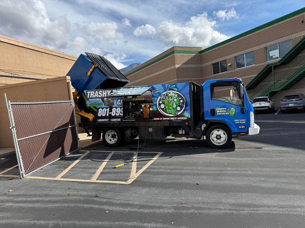 A blue garbage truck lifts a dumpster into its rear hopper inside a parking lot fenced off by a chain-link enclosure.