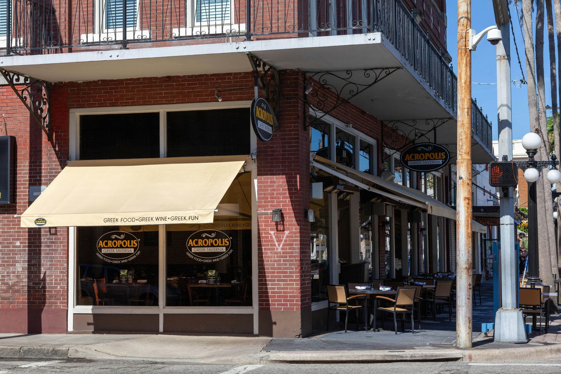 A corner restaurant building with red brick, beige awnings, and outdoor patio seating under a second-story balcony.