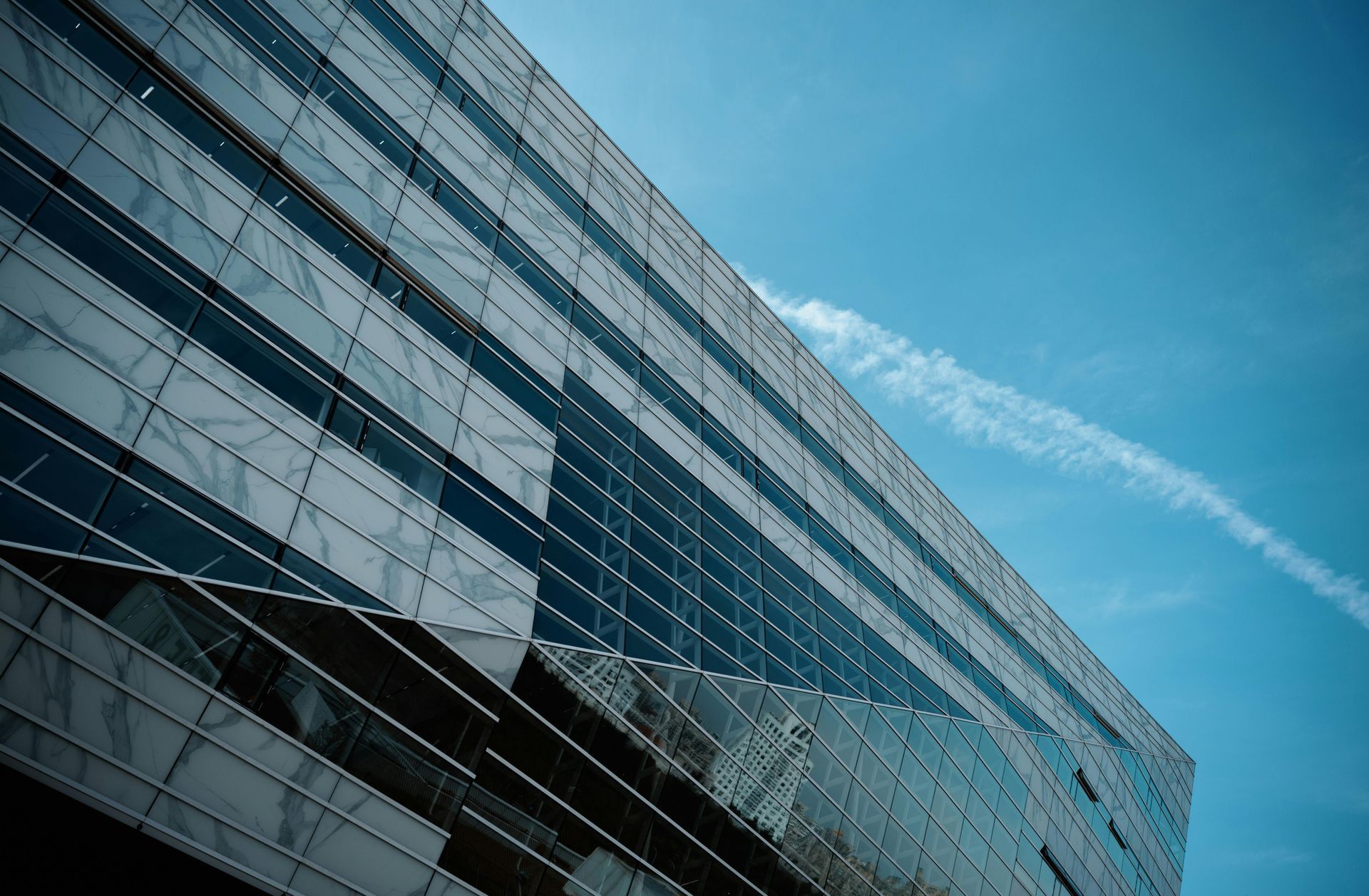 Low-angle shot of a modern glass and steel office building facade against a clear blue sky with a faint vapor trail.