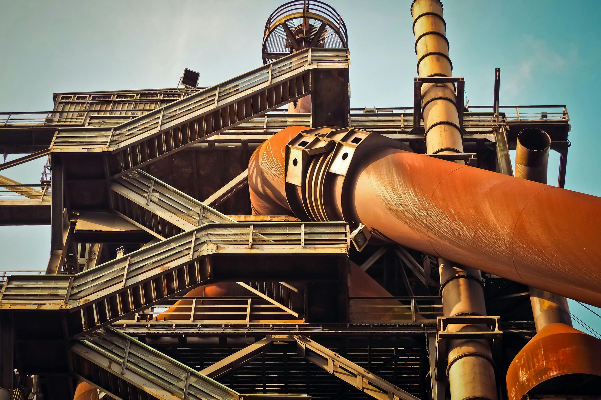 A low-angle view of a weathered, industrial steel structure featuring large rusty pipes and metal stairways against a sky.