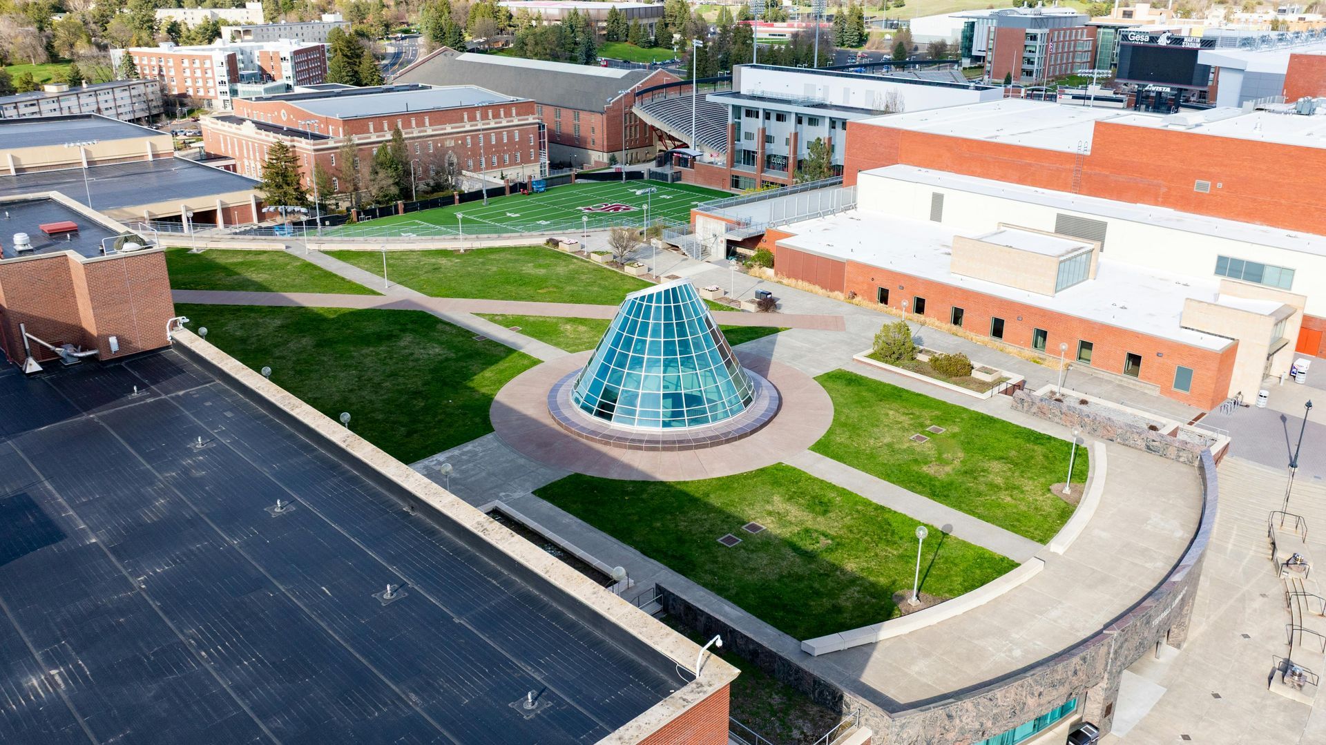 Aerial view of a campus green space featuring a large glass cone structure surrounded by brick buildings and walkways.