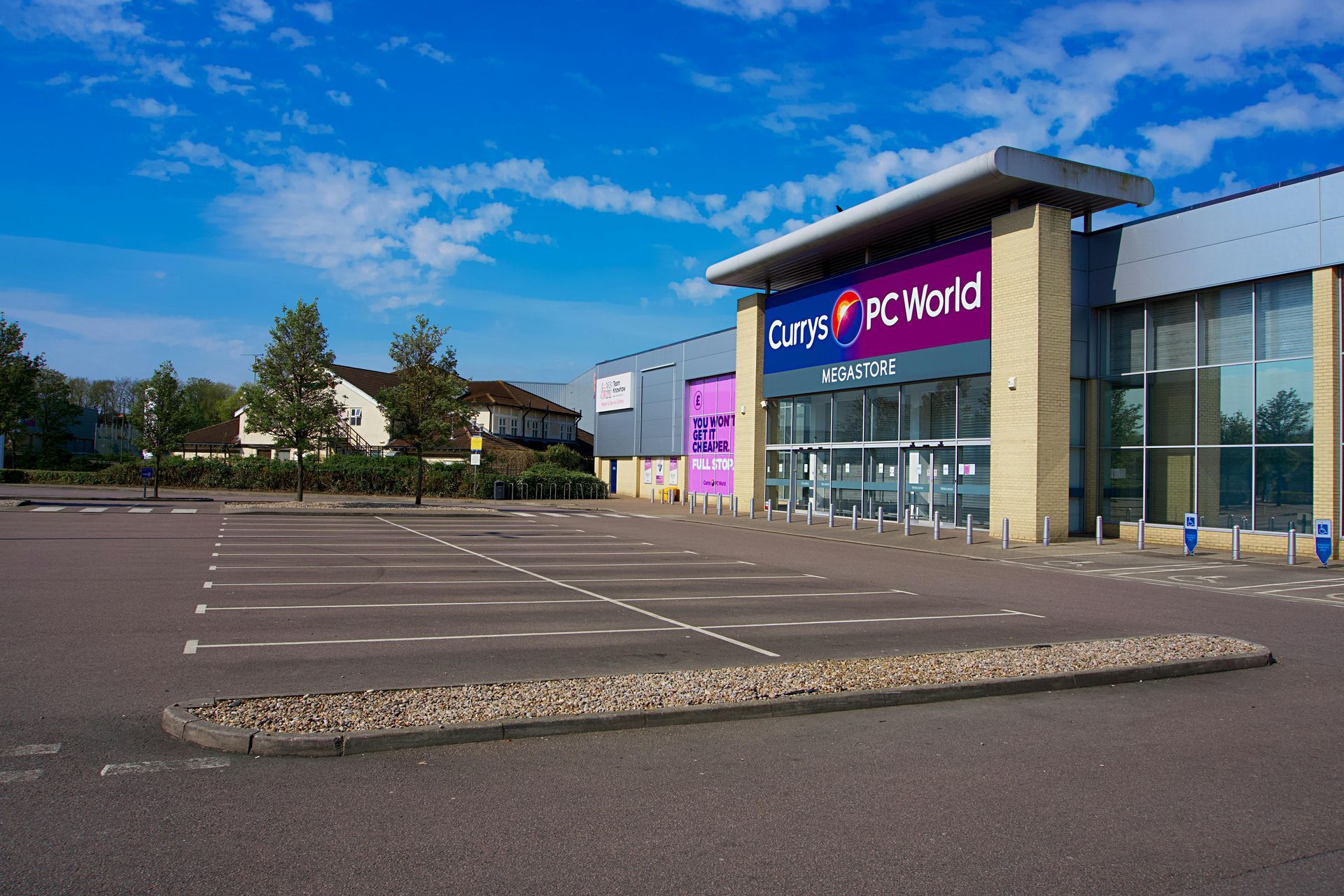 A Currys PC World retail store with a large, empty parking lot under a bright blue sky.