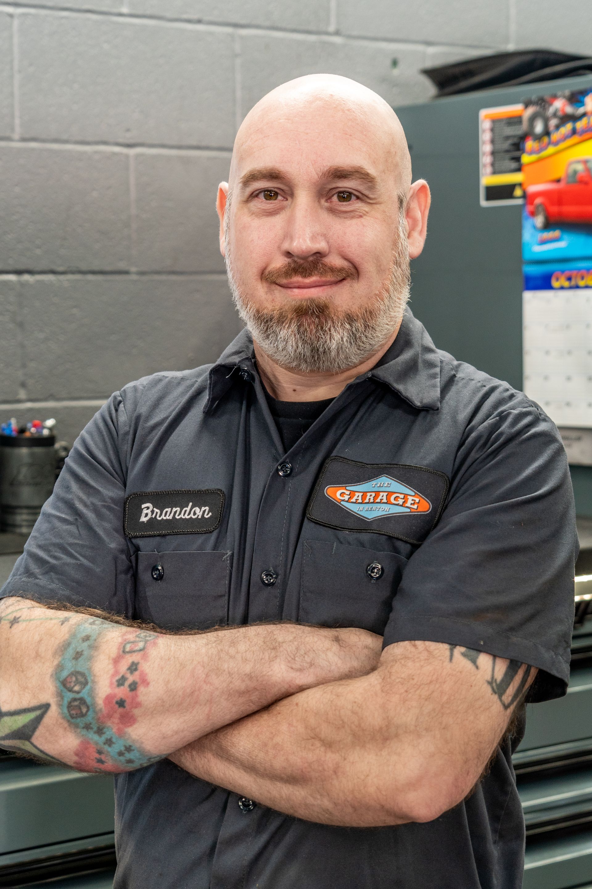 A bald man with a beard and tattoos is standing with his arms crossed in a garage. | The Garage in Renton