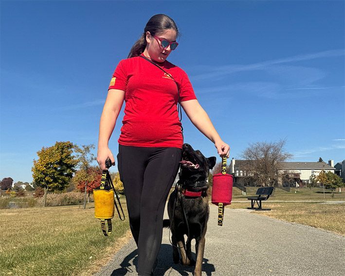Woman in a red shirt walking a black dog on a leash along a sunny park path