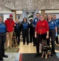 A group of security personnel in blue uniforms and red sweatshirts stand with two working dogs in an airport checkpoint.