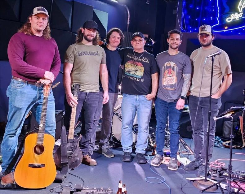 Five people stand on a lit stage with guitars, microphones, and blue string lights in the background.