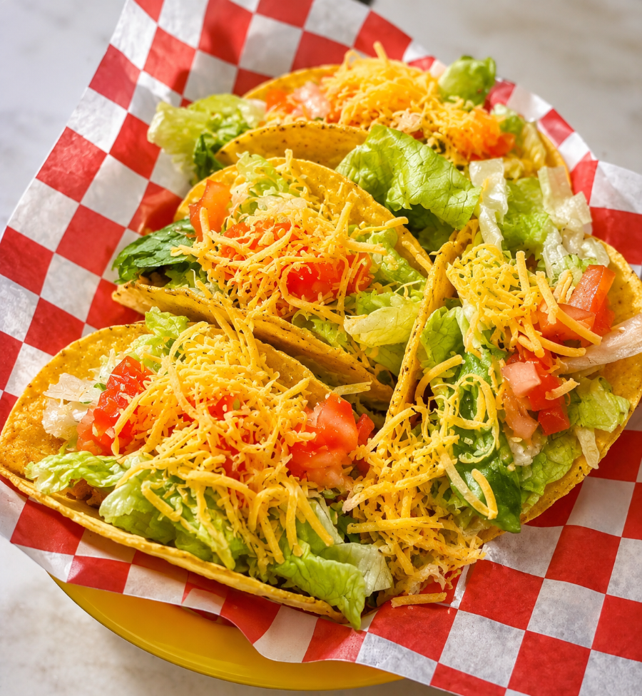 Taco with shredded cheese, lettuce, and diced tomatoes on a red-checkered liner