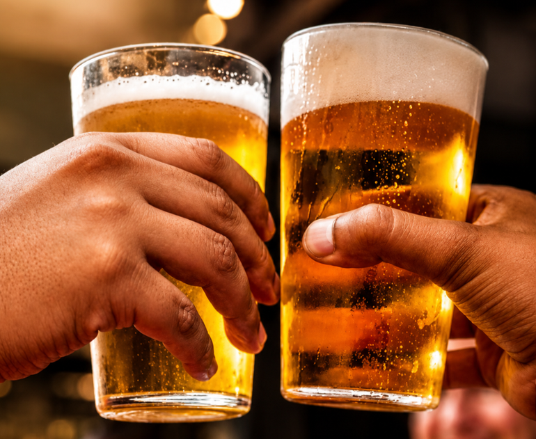 Two hands clink two frothy beer glasses in a dim bar, with blurred people in the background.