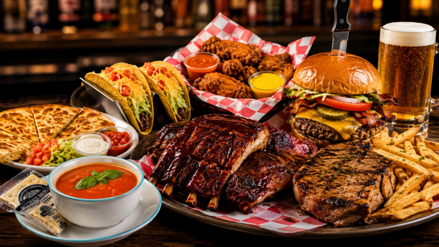 A platter of grilled meats, burgers, tacos, fries, and soups with a beer in a restaurant setting