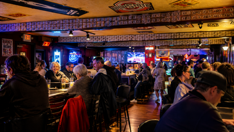 Crowded bar with patrons seated at tables under warm lights and neon signs in a lively pub scene
