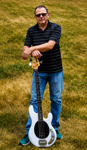 Man standing on a grassy slope holding a guitar beside a blue-and-white guitar leaning against him