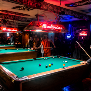 Dimly lit pool hall with a teal billiards table, neon Budweiser signs, and people playing in the background